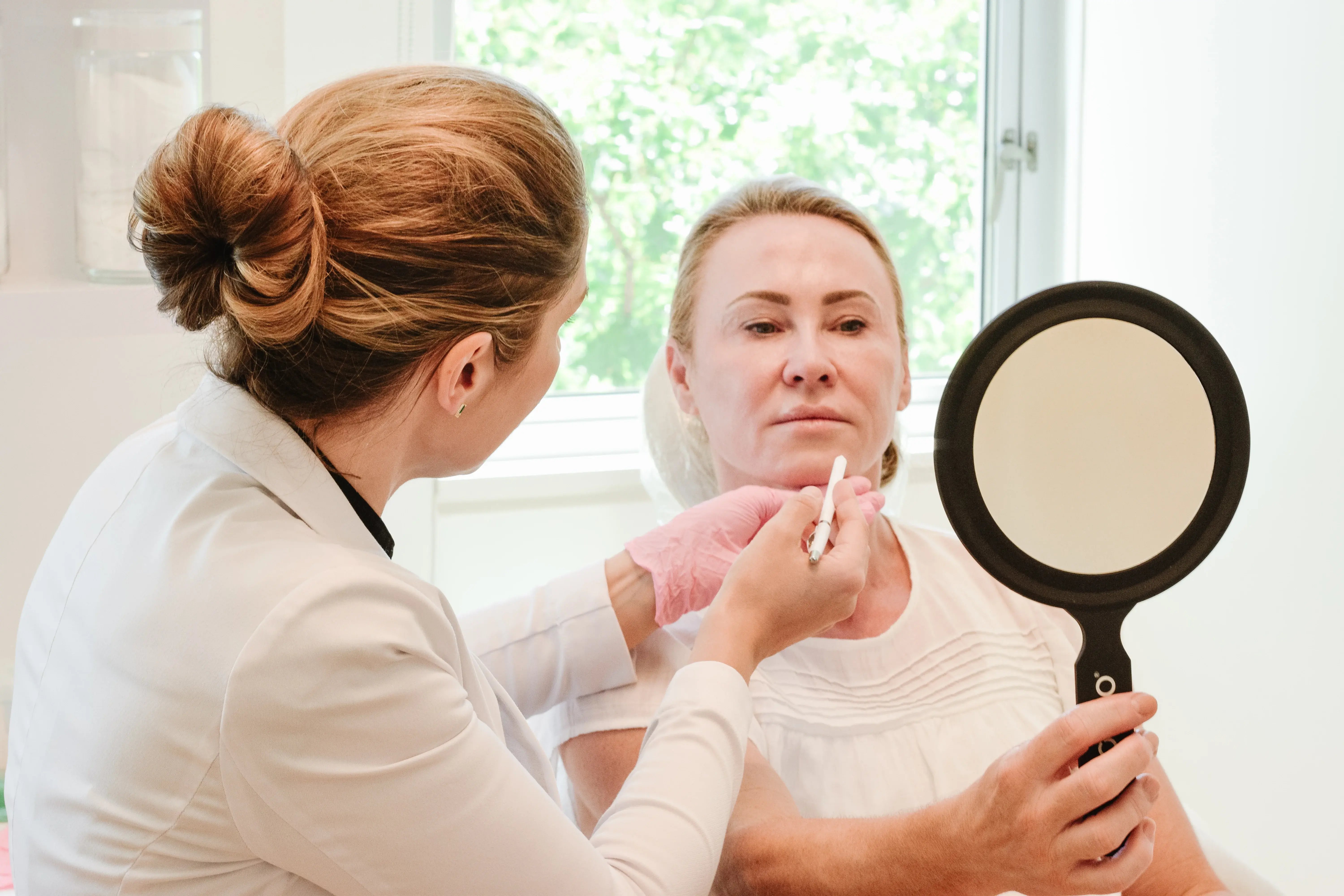 aesthetic-specialist-consulting-with-a-patient-holding-a-mirror-during-a-facial-harmonization-assessment-in-a-bright-treatment-room