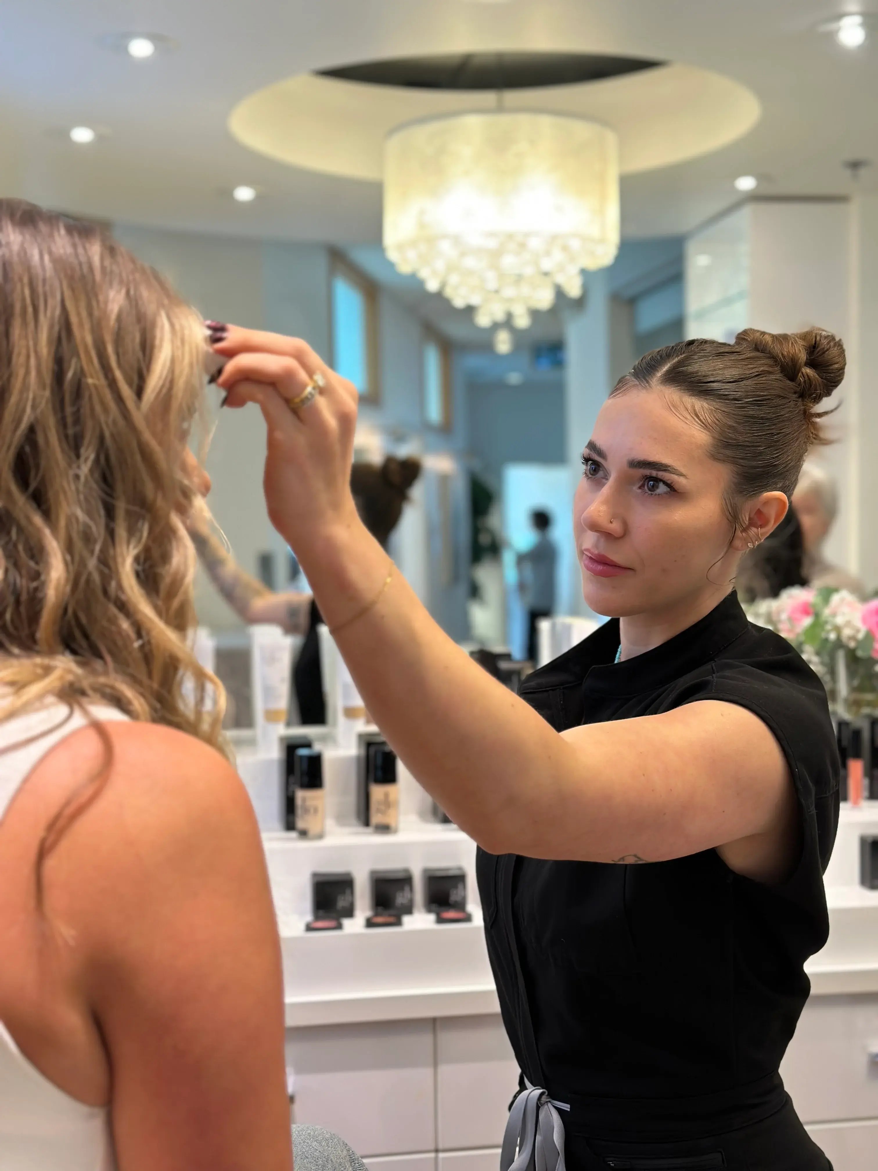 esthetician-applying-product-to-client-in-modern-clinic-with-makeup-display-and-chandelier-in-background