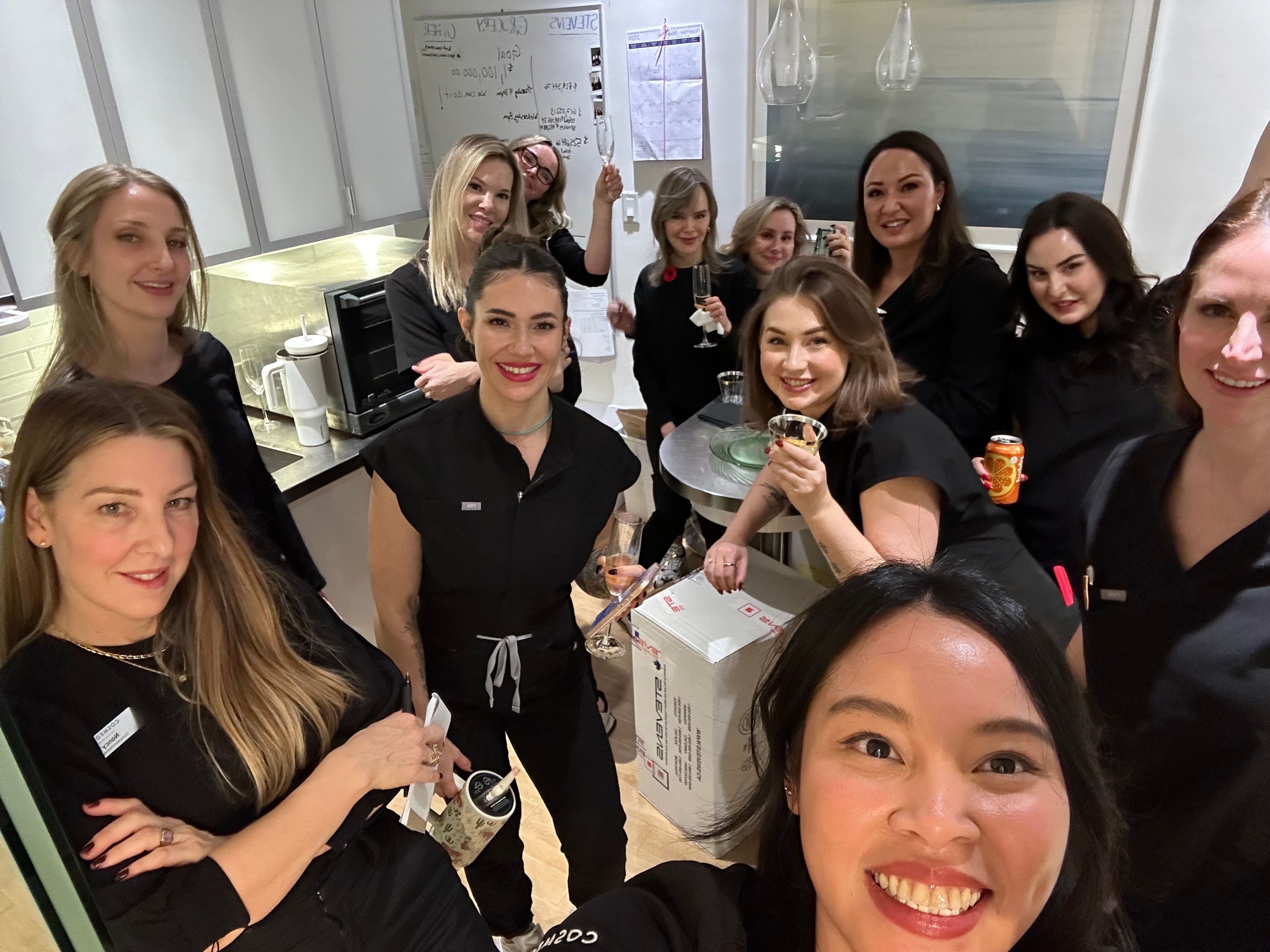 group-of-smiling-staff-in-black-uniforms-holding-drinks-and-celebrating-in-clinic-during-holiday-beauty-sale-event
