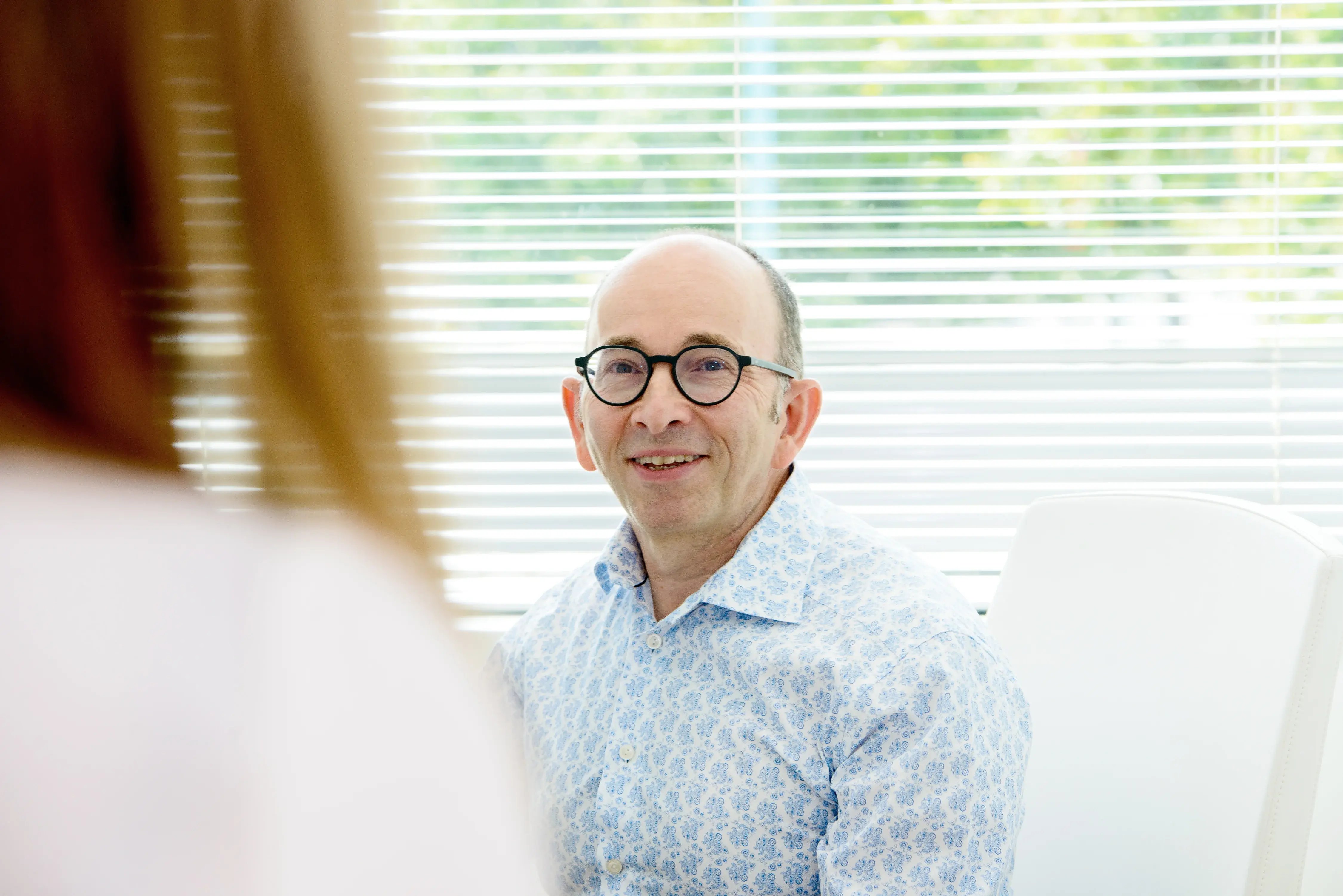 man-wearing-glasses-and-a-light-patterned-shirt-smiling-while-sitting-in-a-bright-office-with-window-blinds-behind-him