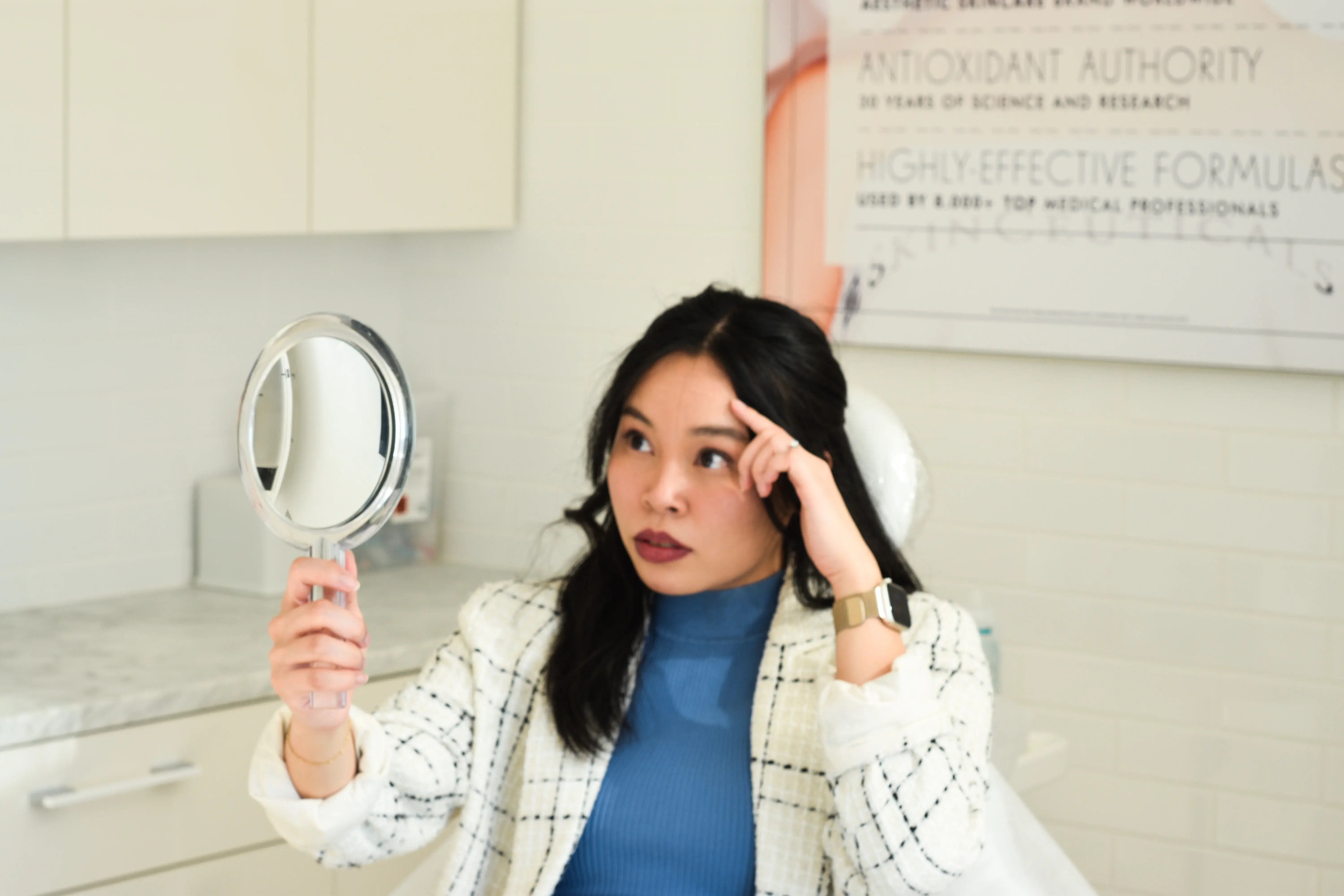 woman-sitting-in-clinic-chair-holding-a-mirror-and-examining-her-forehead-while-thinking-about-melasma-vs-sun-spots-treatment-options