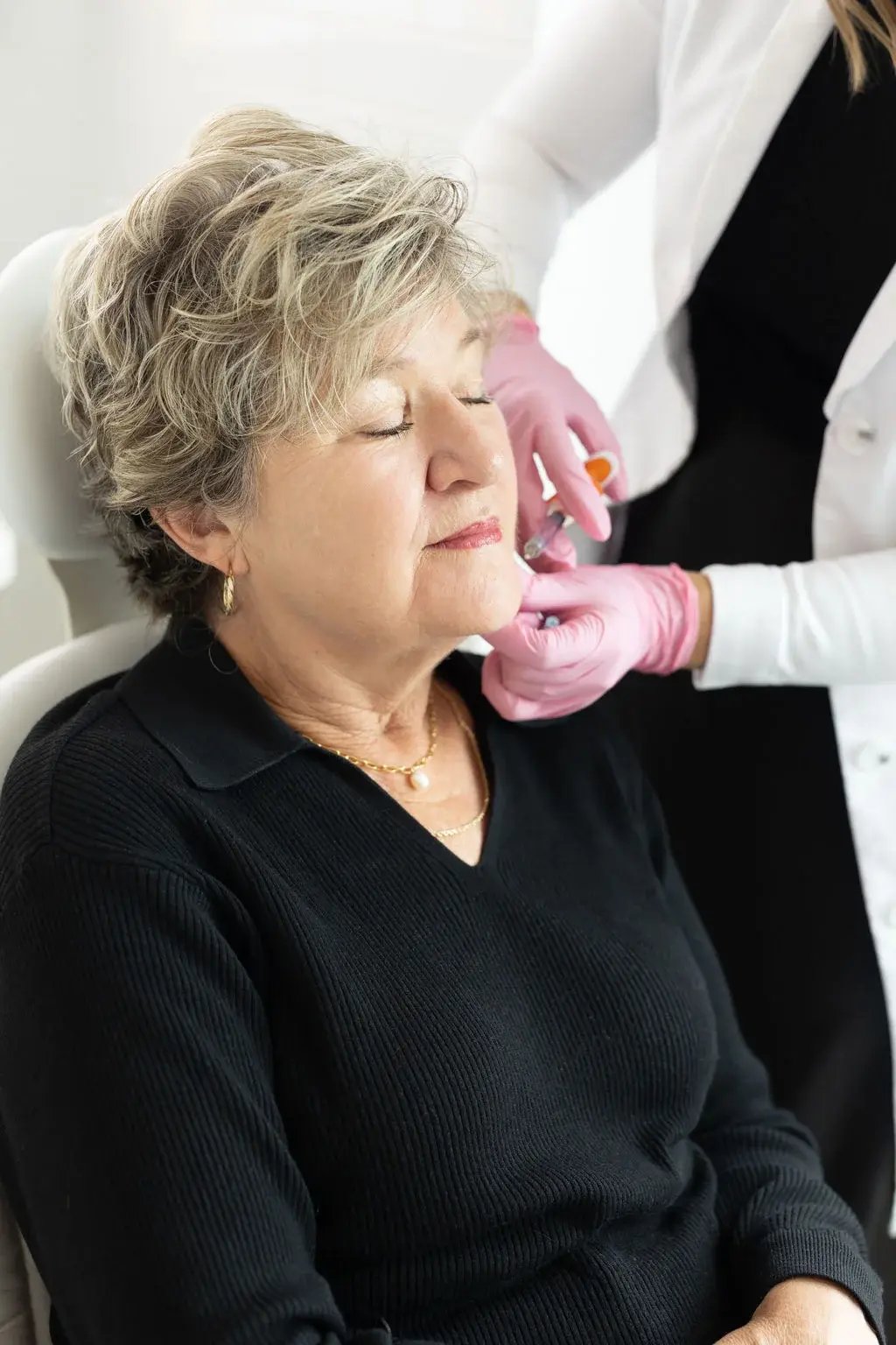 woman-with-closed-eyes-receiving-an-aesthetic-injection-on-her-chin-from-a-clinician-wearing-pink-gloves-in-a-bright-treatment-room